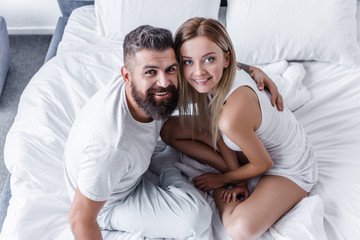overhead view of happy young couple sitting in white bed and looking at camera
