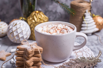 A white cup with hot cocoa with marshmallow stands on a gray table among white and gold Christmas...