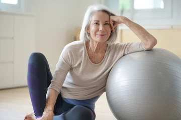 Foto op Canvas  Smiling elderly woman resting on a swiss ball at home © goodluz