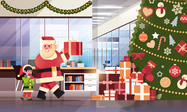santa claus with elf helper put present under decorated fir tree in modern office merry christmas happy new year holidays concept flat horizontal