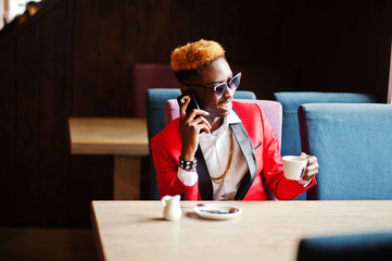 Fashion african american man model at red suit, with highlights hair and sunglasses sitting at cafe with coffee and speaking on mobile phone.
