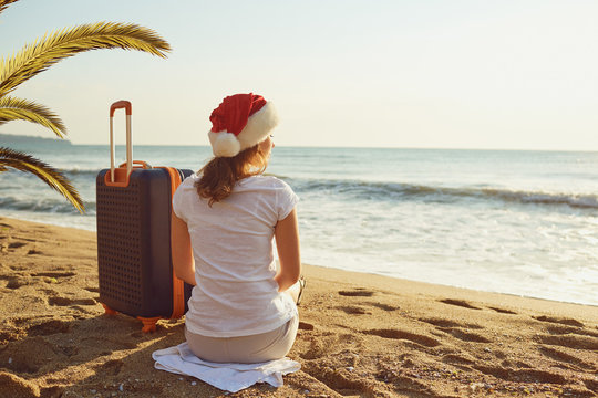 A girl in a Santa Claus hat with a suitcase on the beach on Chri