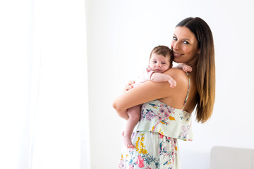 A beautiful young mother standing and holding her baby girl in her arms in front of a white wall.