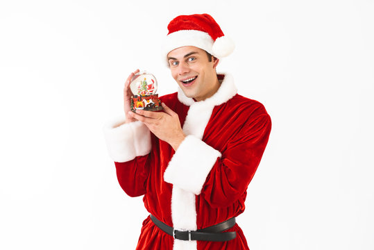 Image of young man 30s in santa claus costume and red hat holding christmas ball, isolated on white background in studio