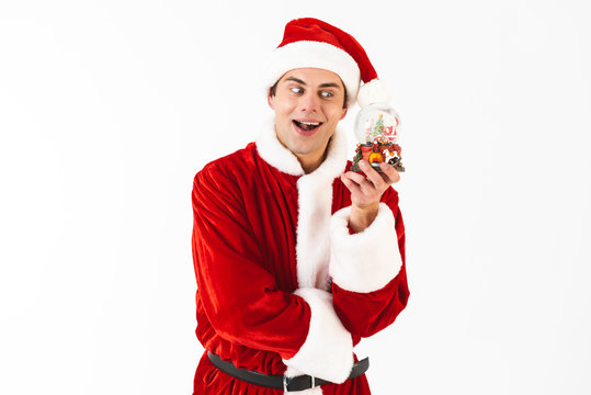 Image of cheerful man 30s in santa claus costume and red hat holding christmas ball, isolated on white background in studio