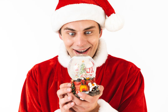 Image of excited man 30s in santa claus costume and red hat holding christmas ball, isolated on white background in studio
