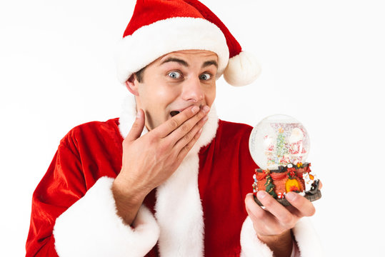 Image of surprised man 30s in santa claus costume and red hat holding christmas ball, isolated on white background in studio