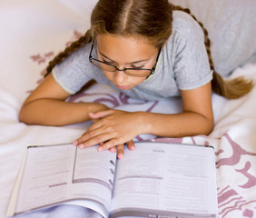 Young girl with eyeglasses reading book on the bed