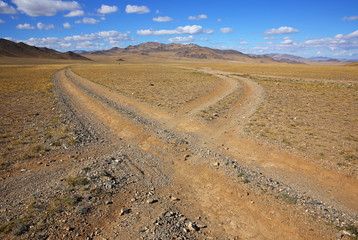 The rural road in the desert behind the mountain range with sky blue and cluods. Central Asia between the Russian Altai and Western Mongolia 