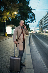 Mature businessman with smartphone and suitcase waiting for a tram in the evening.