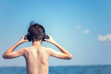 Happy cheerful teen boy on the beach with the headphones running on the wave of the sea on the resort town background. Concept