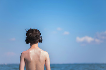 Happy cheerful teen boy on the beach with the headphones running on the wave of the sea on the resort town background. Concept