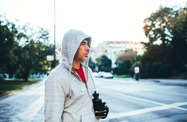 Mature male runner with water bottle standing outdoors in city, resting.