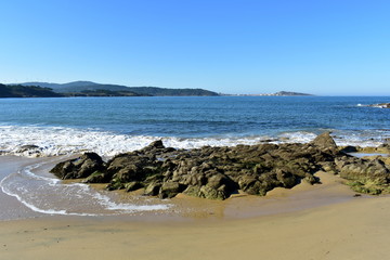 Fototapeta na wymiar Beach in a bay with rocks, wet sand and blue sea with foam. Sunny day, Galicia, Spain.