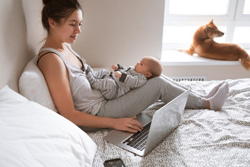 Busy young woman working or study on laptop computer while holding her baby in arms at home
