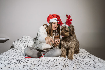 .Young and cheerful woman playing with her nice spanish water dog on Christmas morning, drinking a hot chocolate. Both with christmas costume. Lifestyle.