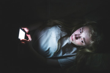 Depressed girl lying down on a couch in the dark while using her smartphone. The light from the...