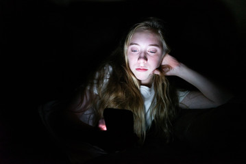 Depressed girl lying down on a couch in the dark while using her smartphone. The light from the screen is illuminating her face.