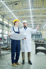 Fototapeta na wymiar Full length portrait of young factory worker discussing work with mature engineer holding plans
