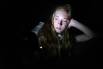 Depressed girl lying down on a couch in the dark while using her smartphone. The light from the screen is illuminating her face.