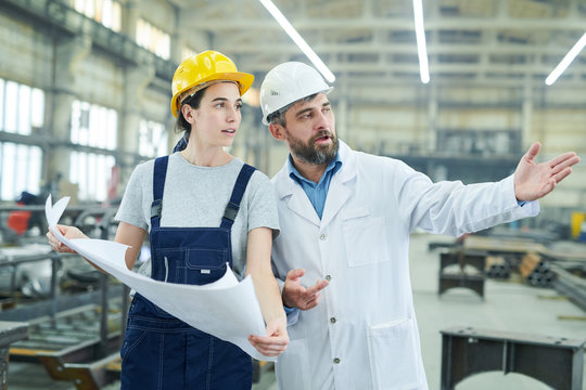 Waist up portrait of female factory worker discussing plans and work with mature engineer in workshop