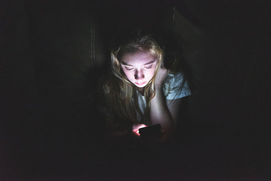 Upset girl sitting in the dark while using her smartphone. The light from the screen is illuminating her face.