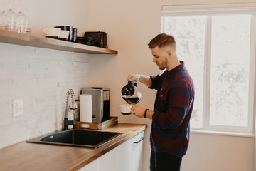 Young Attractive Minimalist Hipster Man in Modern Designed Kitchen Brews, Pours, and Drinks Morning Coffee out of a Cup