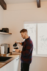 Young Attractive Minimalist Hipster Man in Modern Designed Kitchen Brews, Pours, and Drinks Morning Coffee out of a Cup