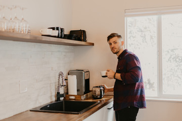 Young Attractive Minimalist Hipster Man in Modern Designed Kitchen Brews, Pours, and Drinks Morning Coffee out of a Cup