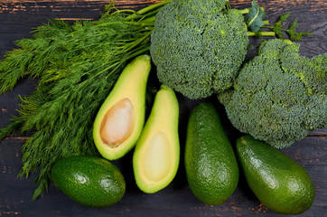 Healthy green food background with raw ingredients for veggie salad or diet dish. Fresh organic vegetables: avocados, dill and broccoli on the dark wooden kitchen table. Top view