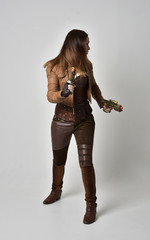 full length portrait of brunette  girl wearing brown leather steampunk outfit. standing pose, holding a gun, on grey studio background.