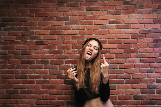 Image of cheeky sporty girl, standing against brick wall with cigarette and showing fuck you gesture