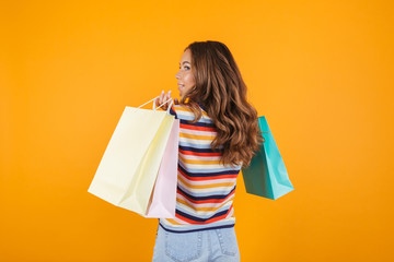 Happy young girl posing over yellow wall background holding shopping bags.