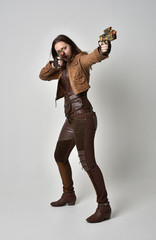 full length portrait of brunette  girl wearing brown leather steampunk outfit. standing pose, holding a gun, on grey studio background.
