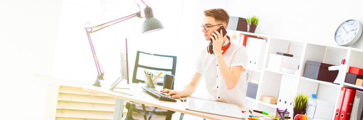 A young man in glasses stands near a computer desk and is talking on the phone. Before him lies a magnetic board and markers. On the neck, the guy's headphones hang. © Ivan Traimak
