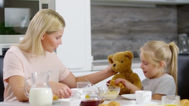 Cute little girl playing with stuffed bear at breakfast: she trying to feed toy with croissant, talking with mother and smiling