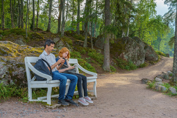 Fototapeta premium Handsome middle-aged man and young pretty lady sitting on bench and using smartphones in summer evening. Tourists on the beautiful landscape background. Monrepos Park, Vyborg, Russia. Travel concept