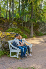 Fototapeta premium Handsome middle-aged man and young pretty lady sitting on bench in summer evening. Tourists on the beautiful landscape background. Monrepos Park, Vyborg, Russia. Travel concept