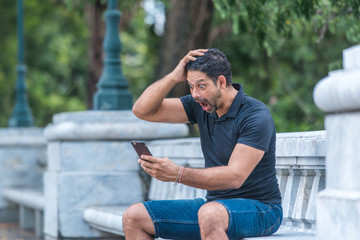 Man holding a smart phone with surprised expression in a park. Emotion and feeling concept