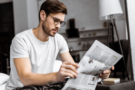 young man in pajamas and eyeglasses sitting on bed and reading newspaper in the morning