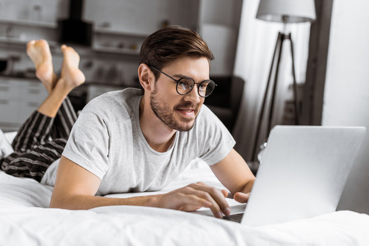 Smiling Young Man In Eyeglasses And Pajamas Using Laptop On Bed