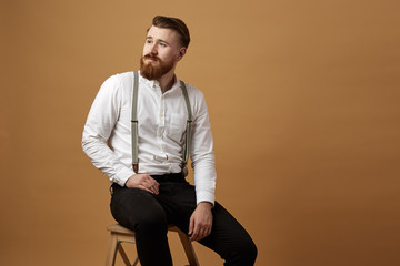 Stylish red-haired man with beard dressed in a white shirt and black trousers with suspender sits...