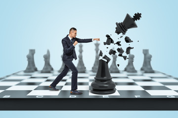 A small businessman stands on a chess board and hits a large black king and destroys it into small pieces.