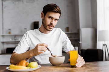 Fototapeta na wymiar handsome young man having cereal with orange juice and looking at camera at home