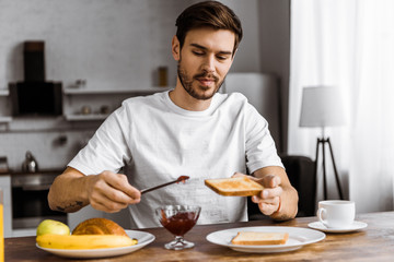 Fototapeta na wymiar smiling young man applying jam onto toast on weekend morning at home