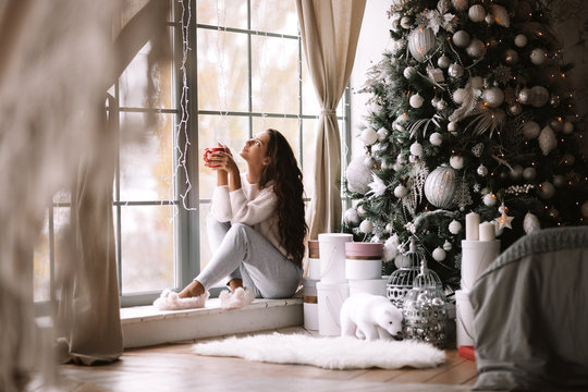 Charming dark-haired girl dressed in pants, sweater and warm slippers holds a red cup sitting on the windowsill of a panoramic window in the room next to the New Year tree, gifts and candles