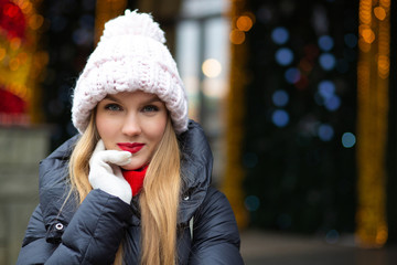 Gorgeous blonde girl with red lipstick wearing knitted cap posing at the street over a garlands background with bokeh