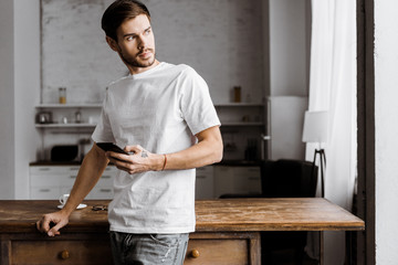 handsome young man using smartphone and looking away on kitchen at home