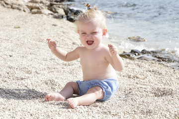 Candid photo of adorable blonde baby on the pebble beach near the sea, summer vacation on the seaside