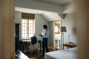 Middle-aged Asian businessman standing at hotel room and working on his tablet.